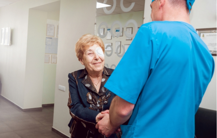 An older man wearing an eye shield on her left eye shaking hands with her ophthalmologist smiling and thanking him for completing cataract surgery