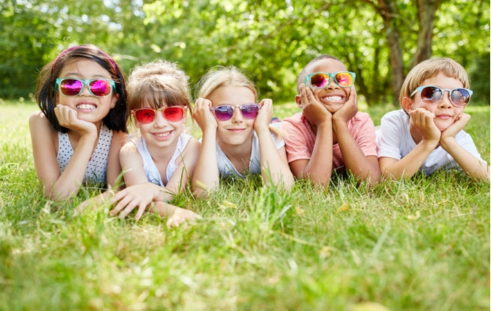 A group of five kids laying on grass all beside each other on a summer day, wearing colourful sunglasses to protect their eyes from UV rays