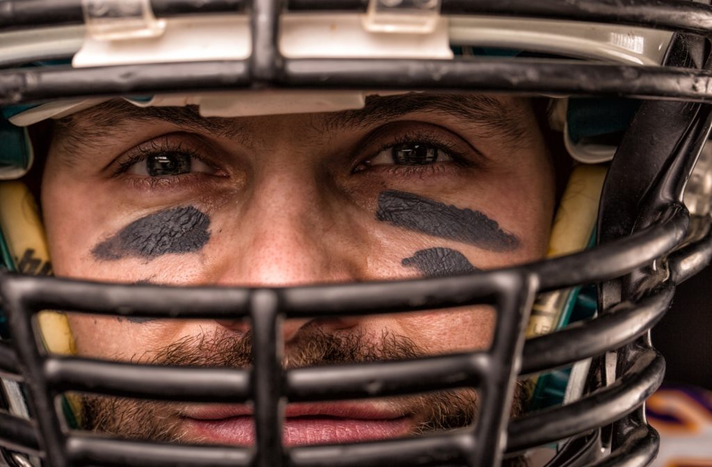 A close up of a male football player's face, wearing a helmet with a black cage and eye black applied underneath his eyes