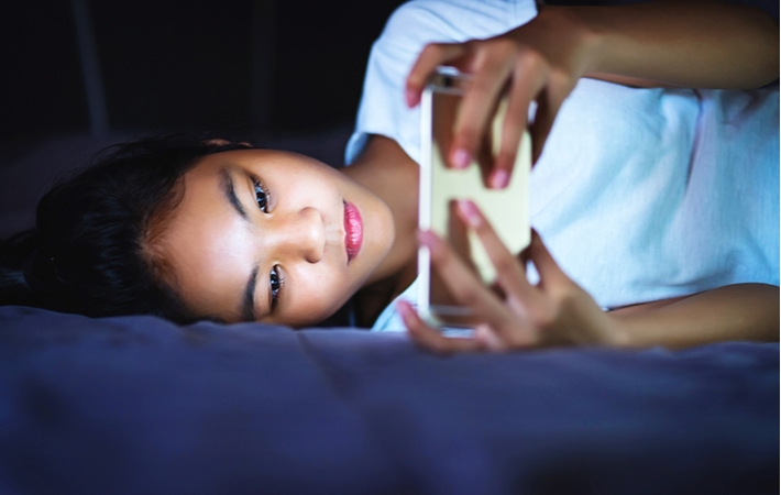 A girl laying down on her bed, using a smartphone that is omitting blue light in her dark room