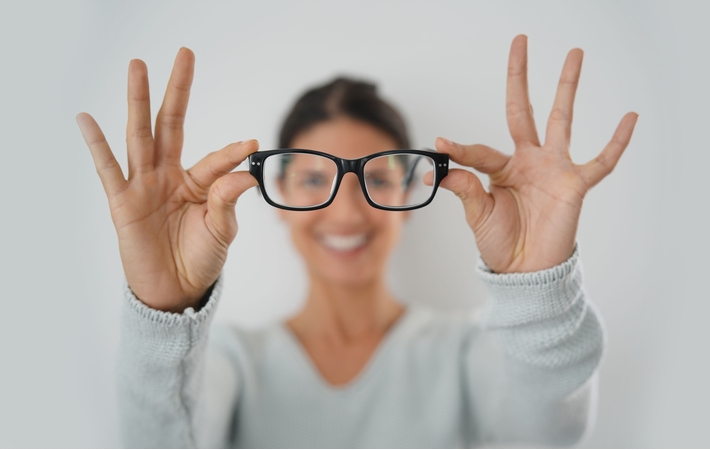 A woman stretching both of her arms forward, holding a pair of black glasses with the rest of her body blurred in the background