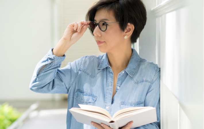 A woman standing, holding a book but looking to the side with progressive lenses, giving her the ability to see clearly up close and into the distance