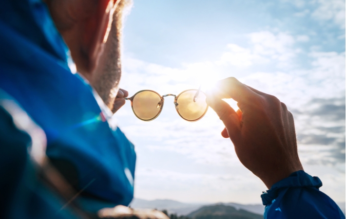 An over the shoulder view of a man wearing hiking gear, holding his polarized sunglasses up to the sunlight