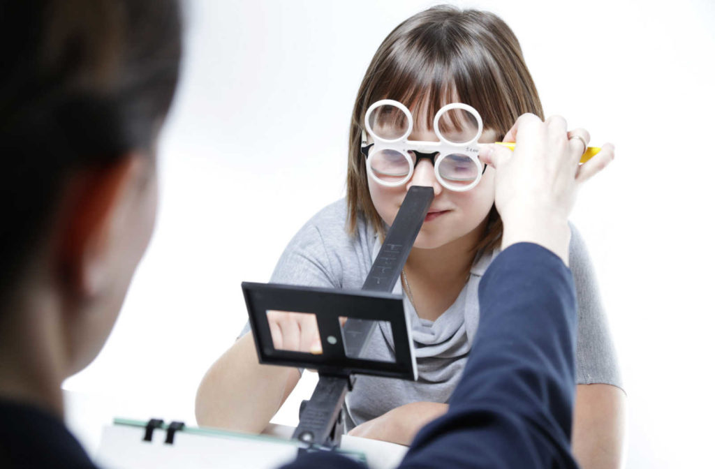 A girl looking through prism lenses onto an object on a scale with her eye doctor in white background.