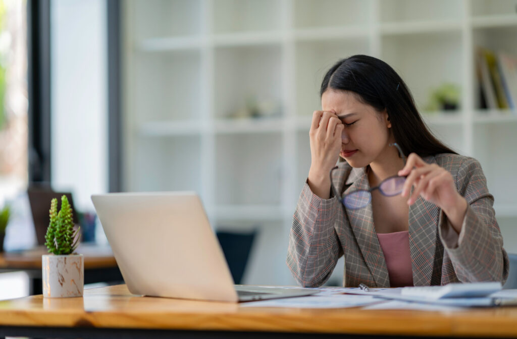 A young woman sitting at a desk in an office with her laptop and holding her glasses in her left hand as she rubs her eyes