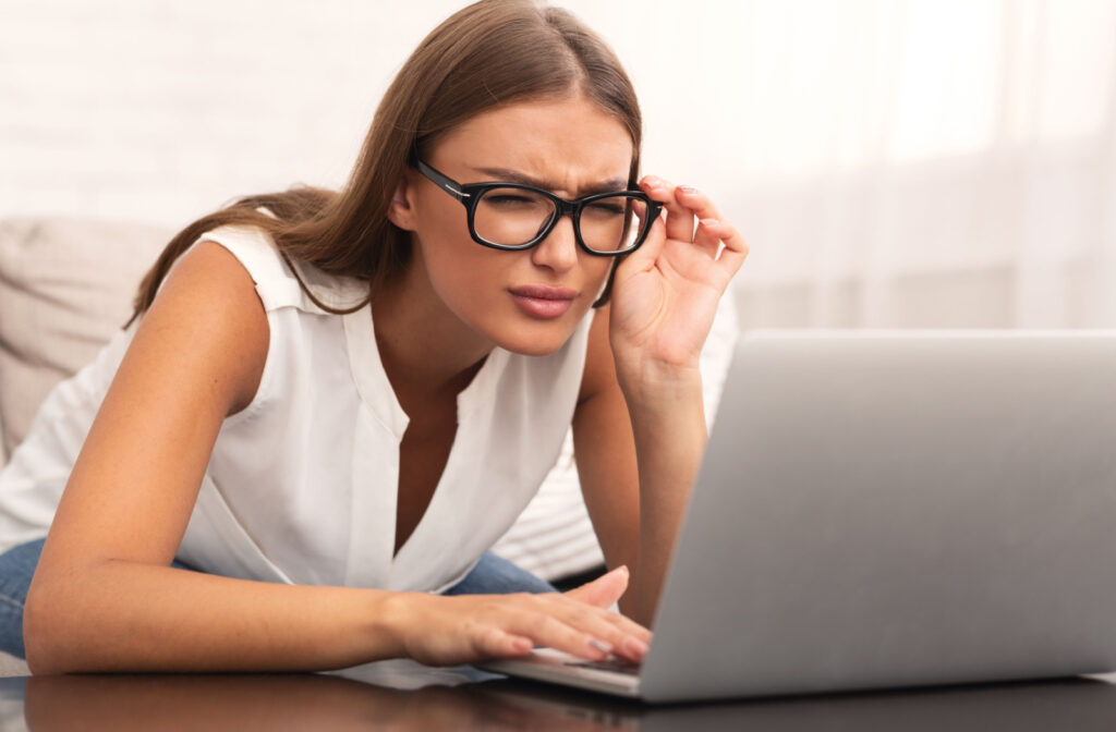 A woman sitting in front of a laptop and adjusting her glasses as she squints to see