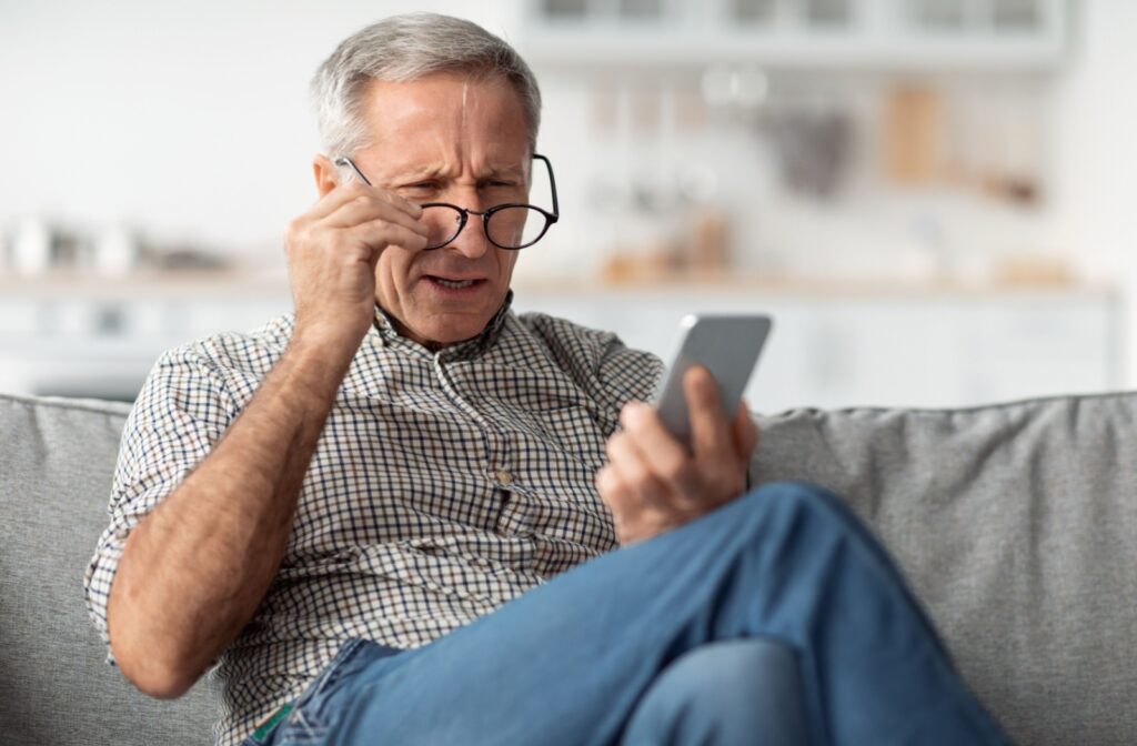 A senior man wearing glasses and squinting his eyes to see more clearly while holding a phone in his left hand.