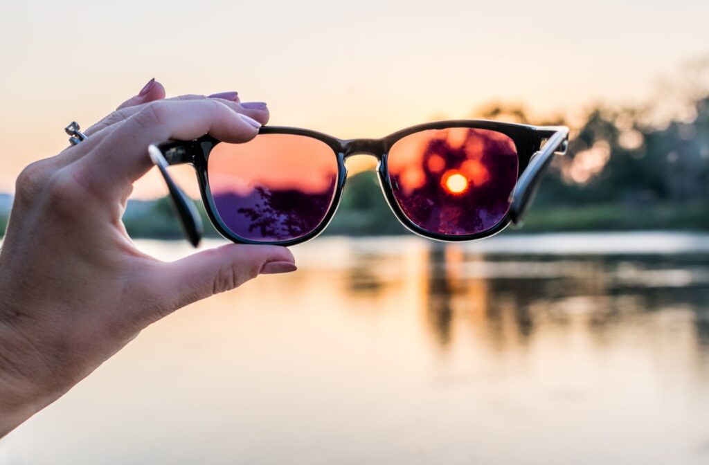 A woman's left hand holding a pair of pink-tinted sunglasses up to a view of a sunset over a lake.