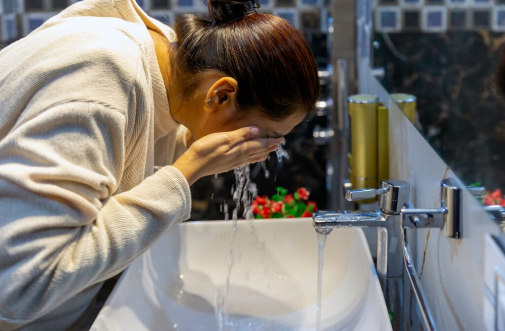 A woman washing her face at a sink, splashing water over the eyes, as part of an eye care cleansing routine.